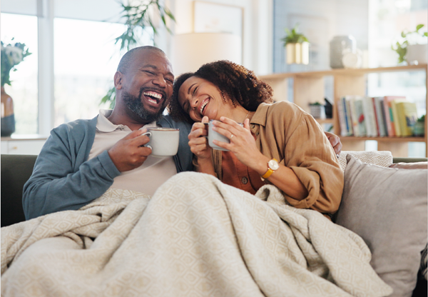 Couple sitting on the couch with a blanket laughing and drinking coffee
